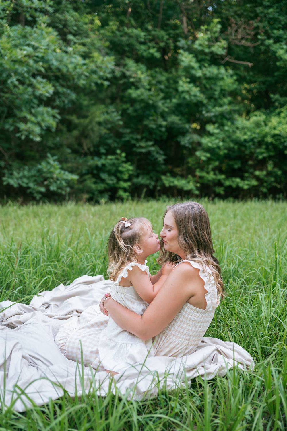 Mother and toddler daughter sharing a sweet kiss while sitting in tall grass in a meadow Asheboro NC