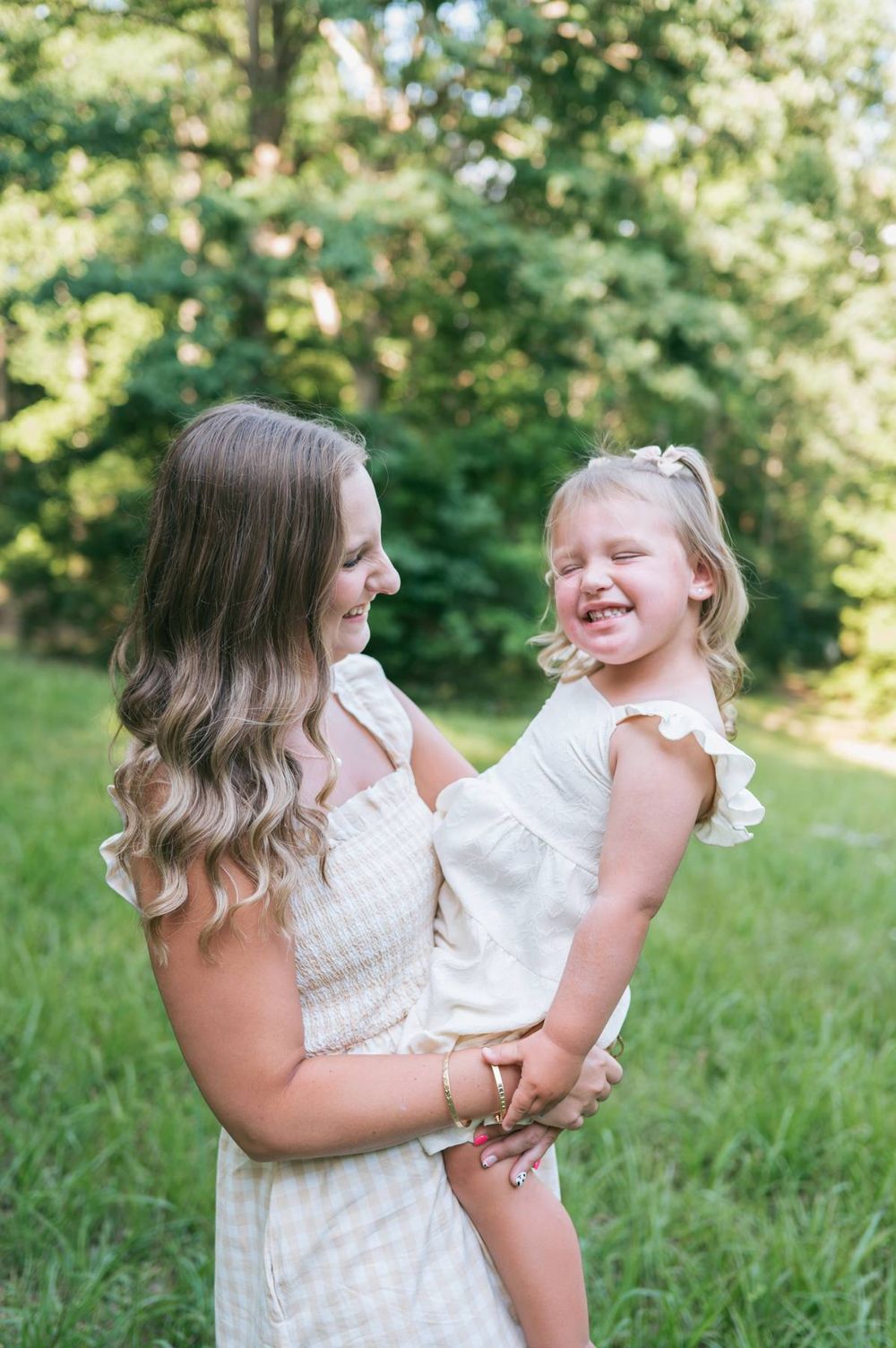 Mother holding her laughing toddler daughter in a sunny green meadow with trees in background in North Carolina