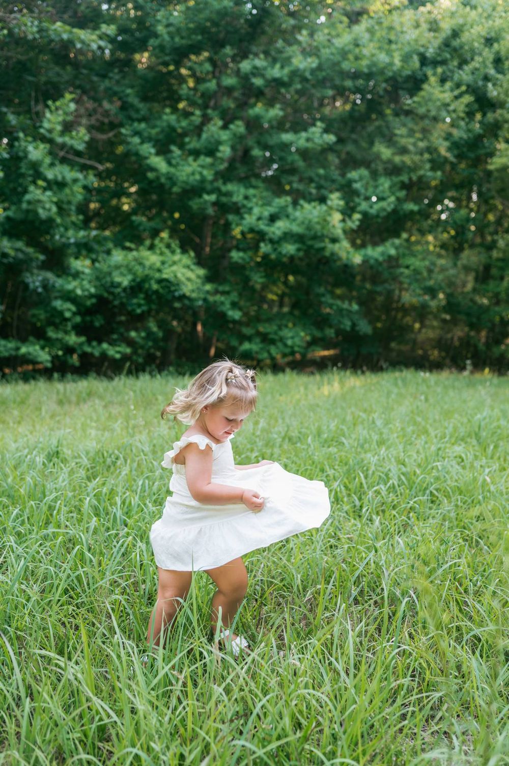 Toddler girl in white dress twirling in a lush green meadow surrounded by trees High Point NC