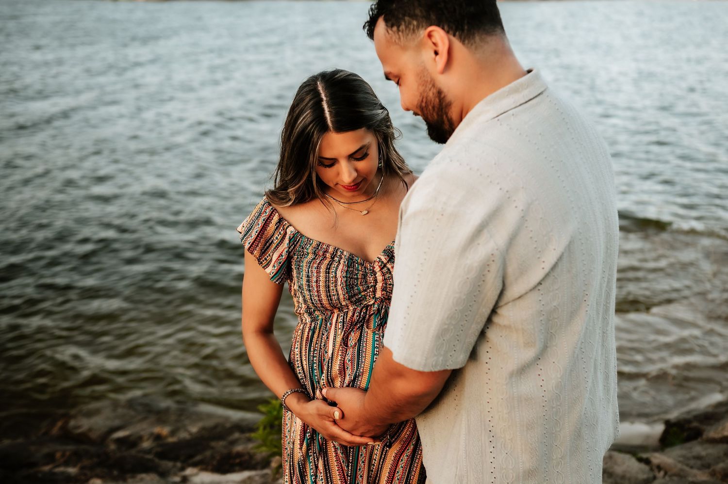 Expectant couple tenderly cradling baby bump together near water during golden hour maternity shoot.