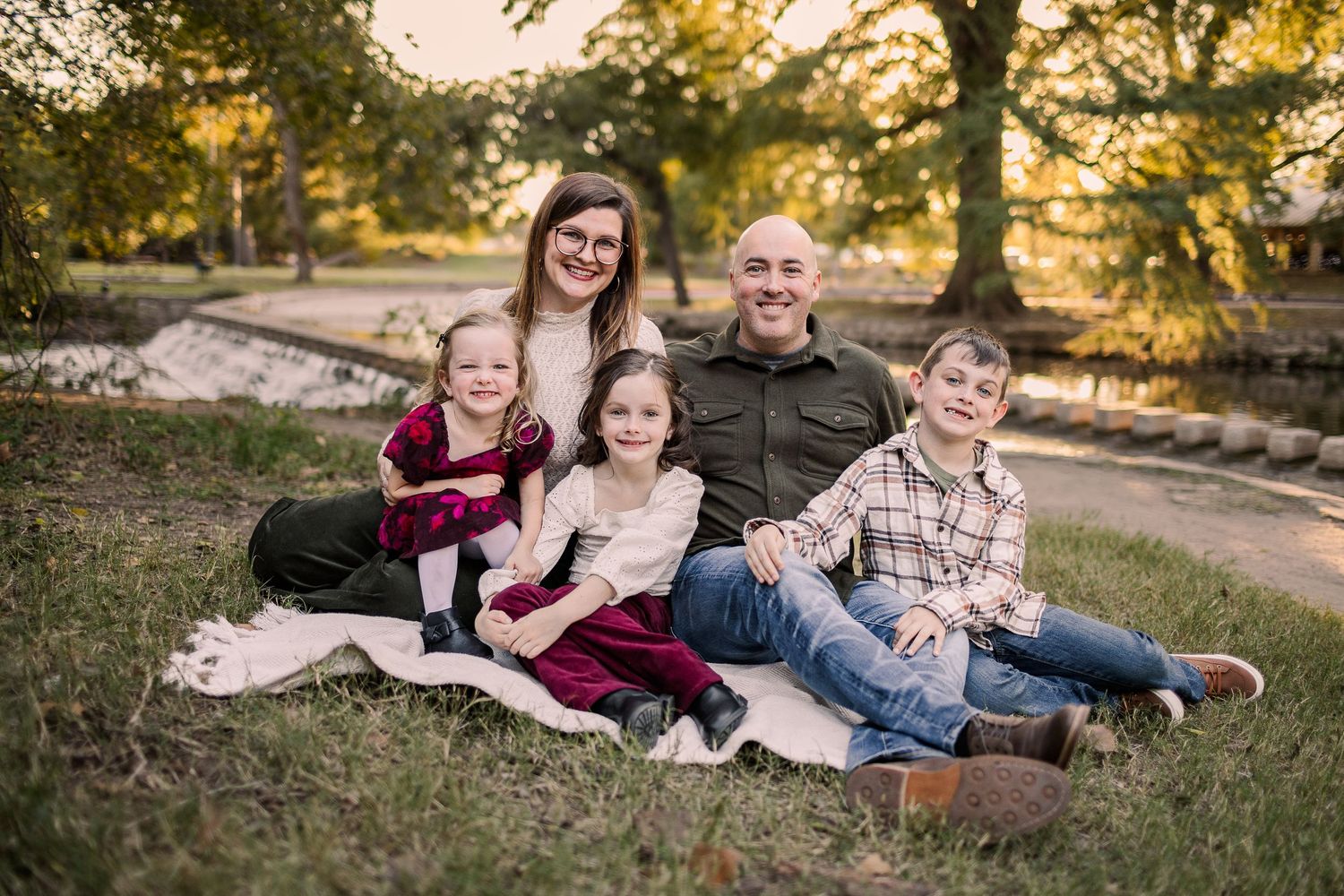 Family of five smiling outdoors near a pond, surrounded by lush green trees during a warm autumn evening.