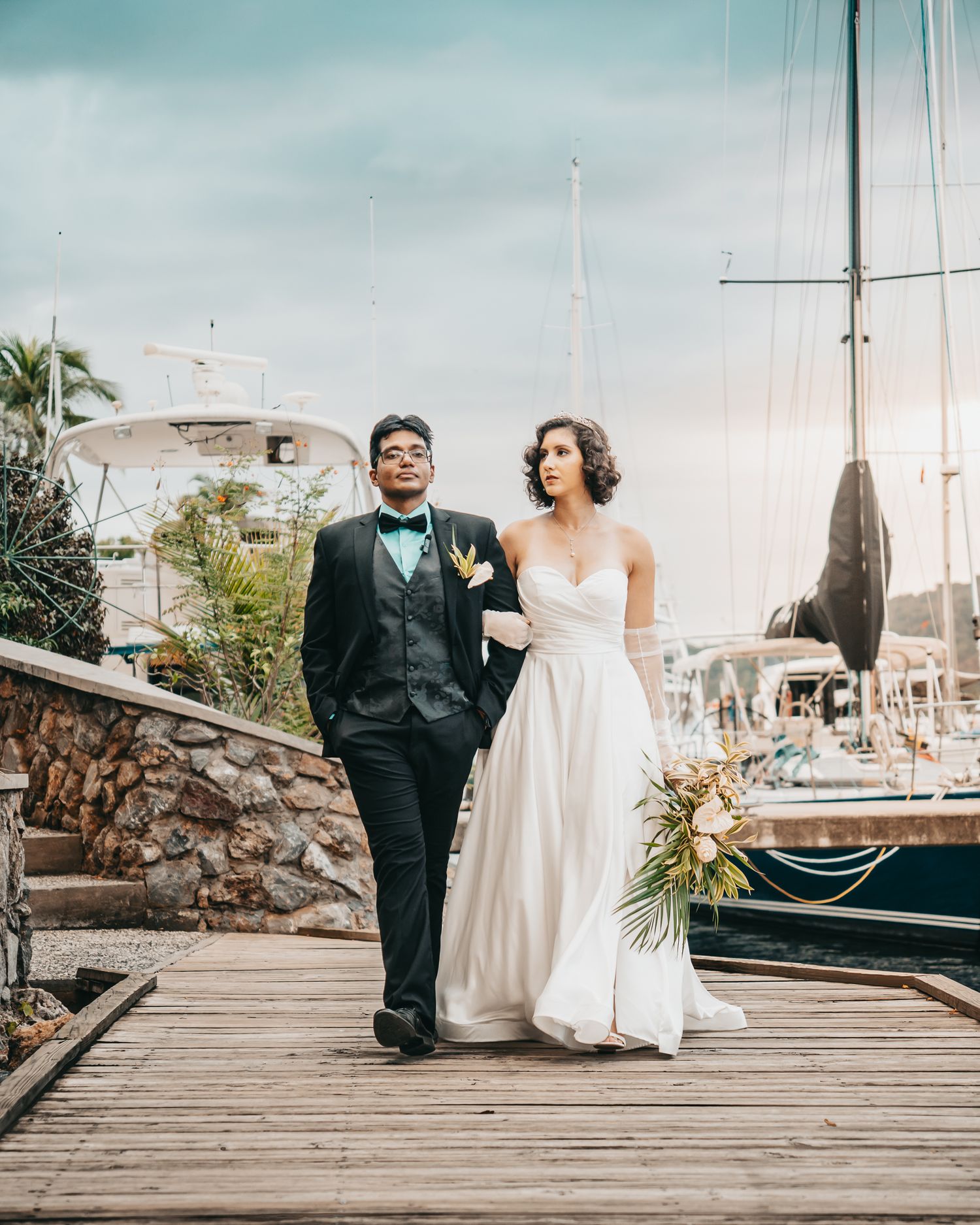 Bride and groom walk hand-in-hand on a marina dock, with sailboats in the background on their wedding day.