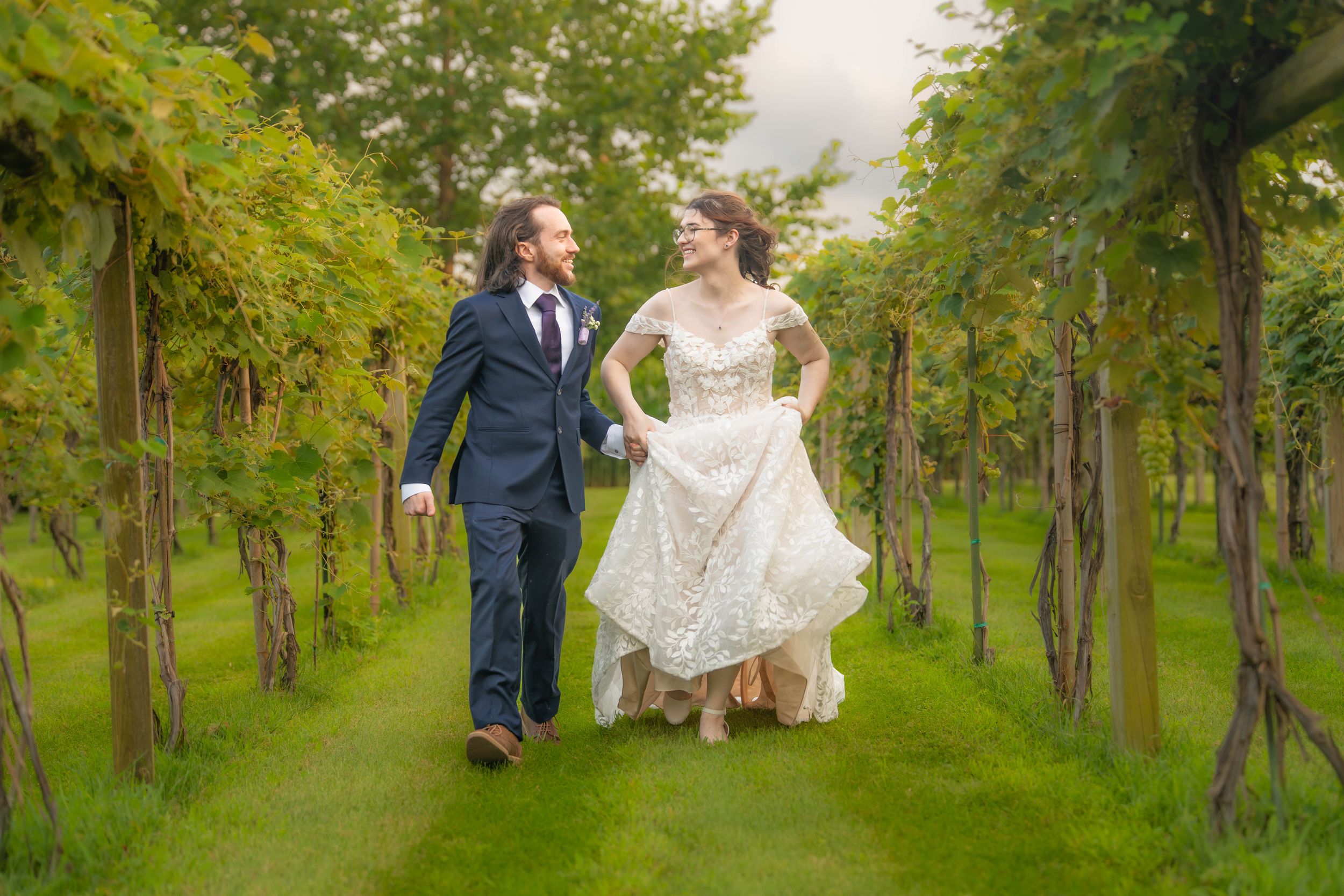 Bride and groom walking hand-in-hand through lush vineyard rows, bride in lace gown, groom in navy suit. Quad Cities Wedding Photographer