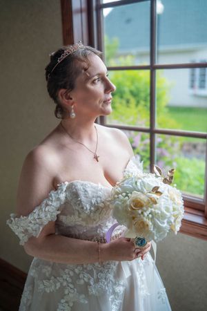 Bride in lace off-shoulder gown holding white rose bouquet, wearing tiara, gazing out window on wedding day. Quad Cities Wedding Photography