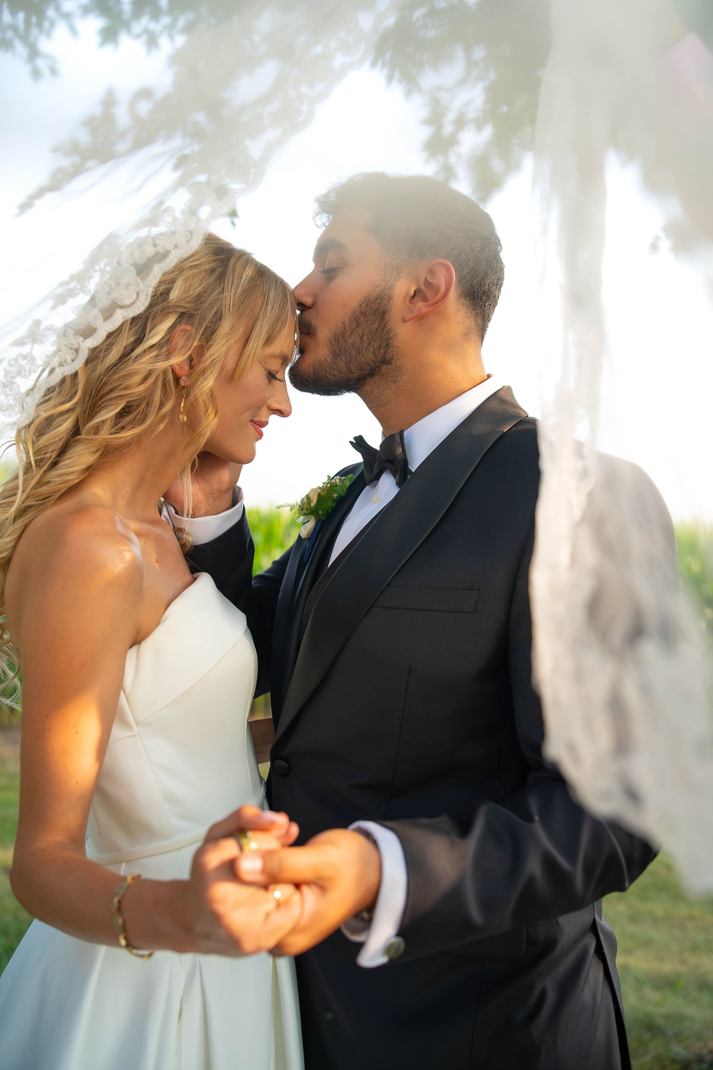 A bride and groom share a romantic kiss outdoors, holding hands, dressed in a white gown and black tuxedo.  Quad Cities Wedding Photographer