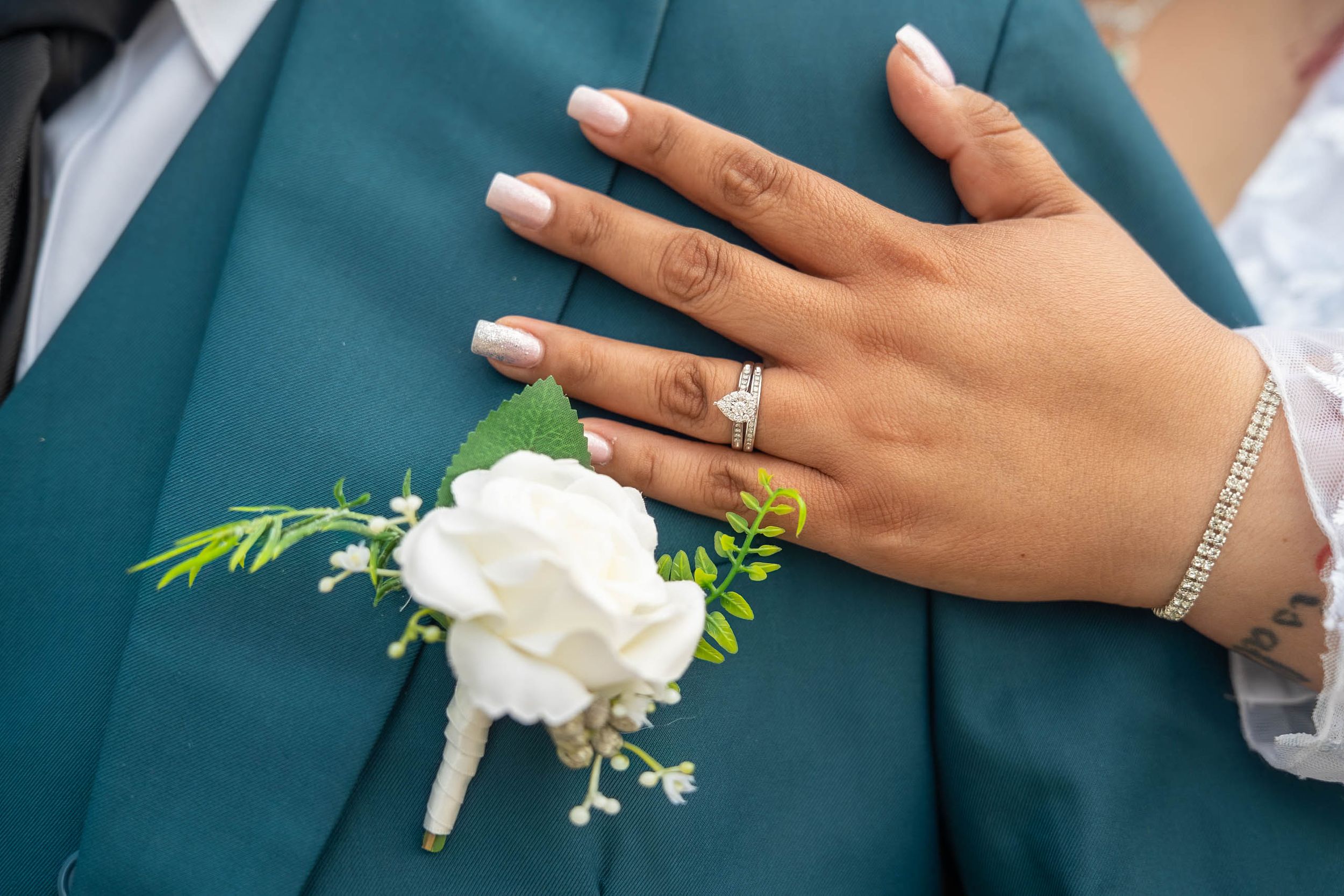 Bride's hand with diamond ring and bracelet resting on groom's teal suit with white rose boutonniere.