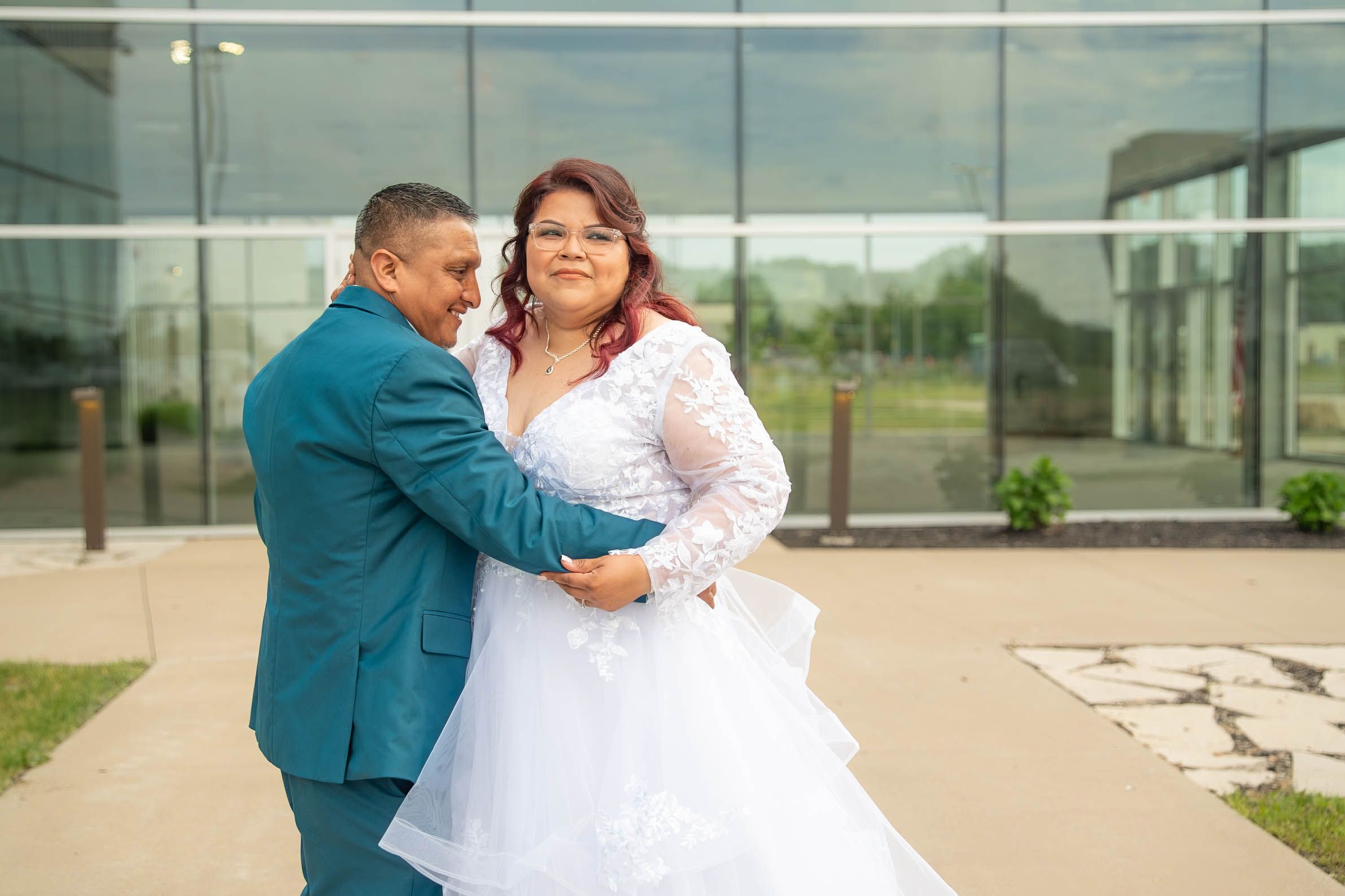 A couple poses outdoors in wedding attire, the groom in a teal suit and bride in a white lace gown.