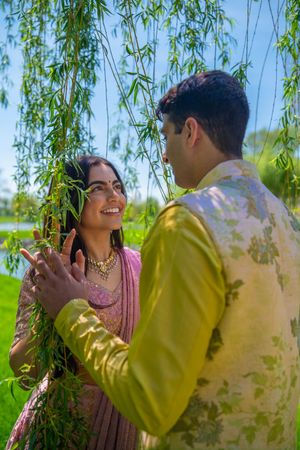 Indian couple in traditional attire smiling under willow tree, woman in pink saree and man in green sherwani.  Quad Cities Wedding Photo