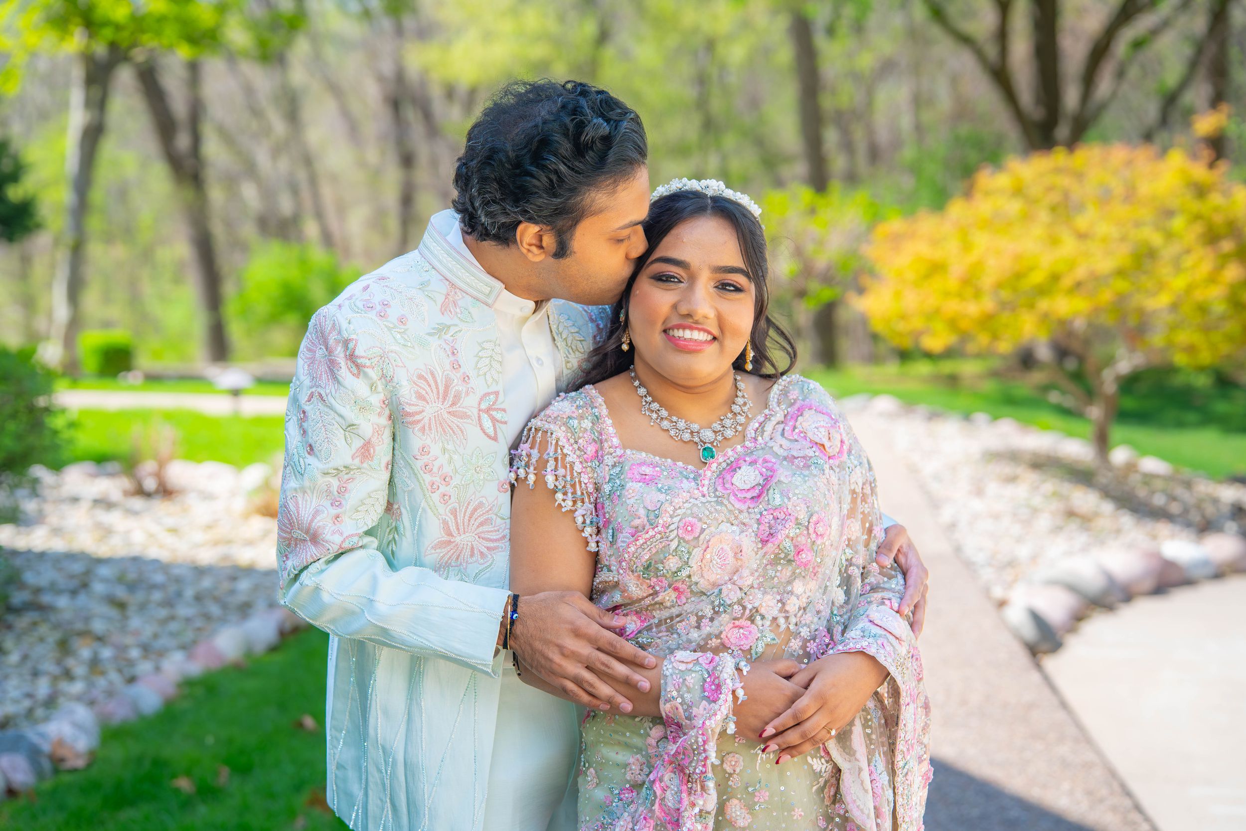 South Asian couple in traditional attire embracing outdoors, man kissing woman's cheek, lush green park background.  Quad Cities Wedding