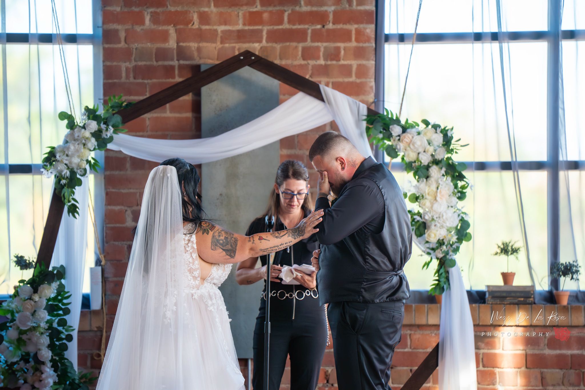 Bride and groom exchange vows at indoor wedding ceremony beneath floral arch with white draping and brick wall backdrop.  QCA Wedding Photo