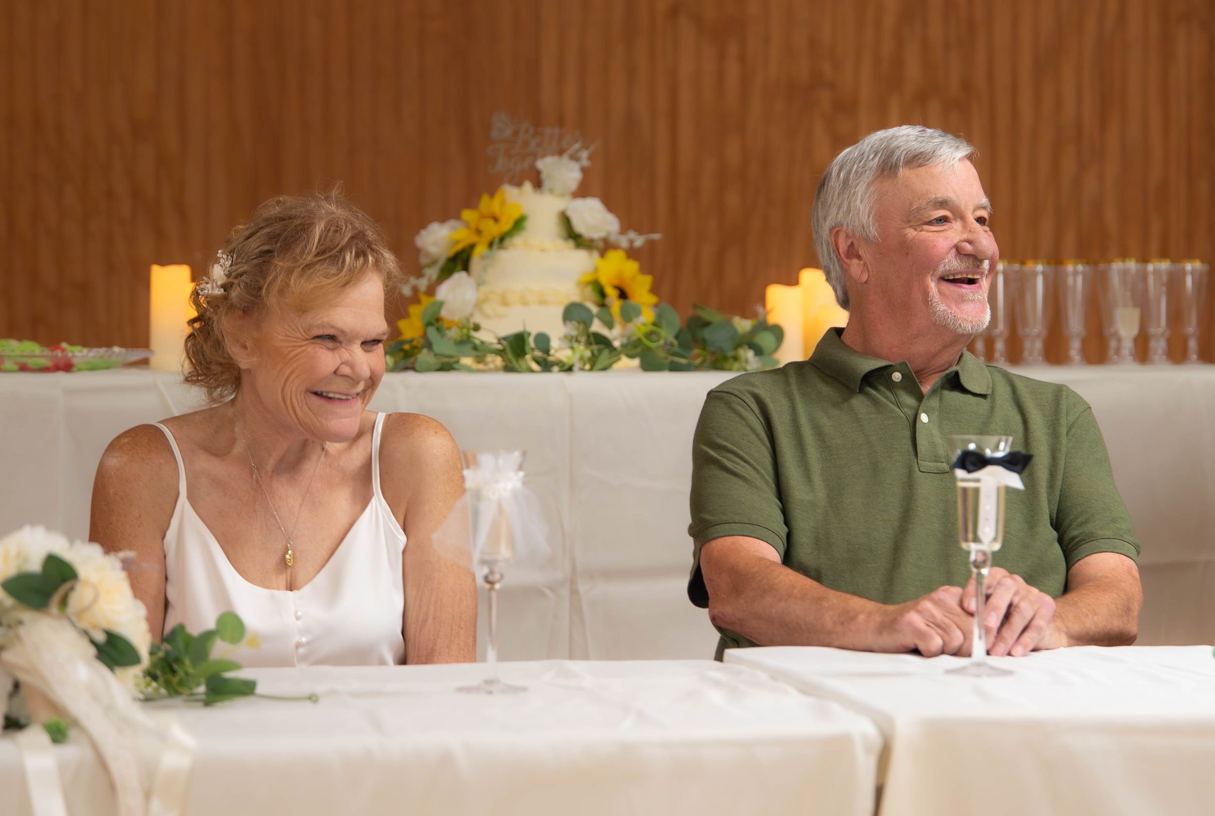 Smiling older couple sitting at a wedding reception table with a white cake and floral arrangement behind them.  Quad Cities Wedding Photo
