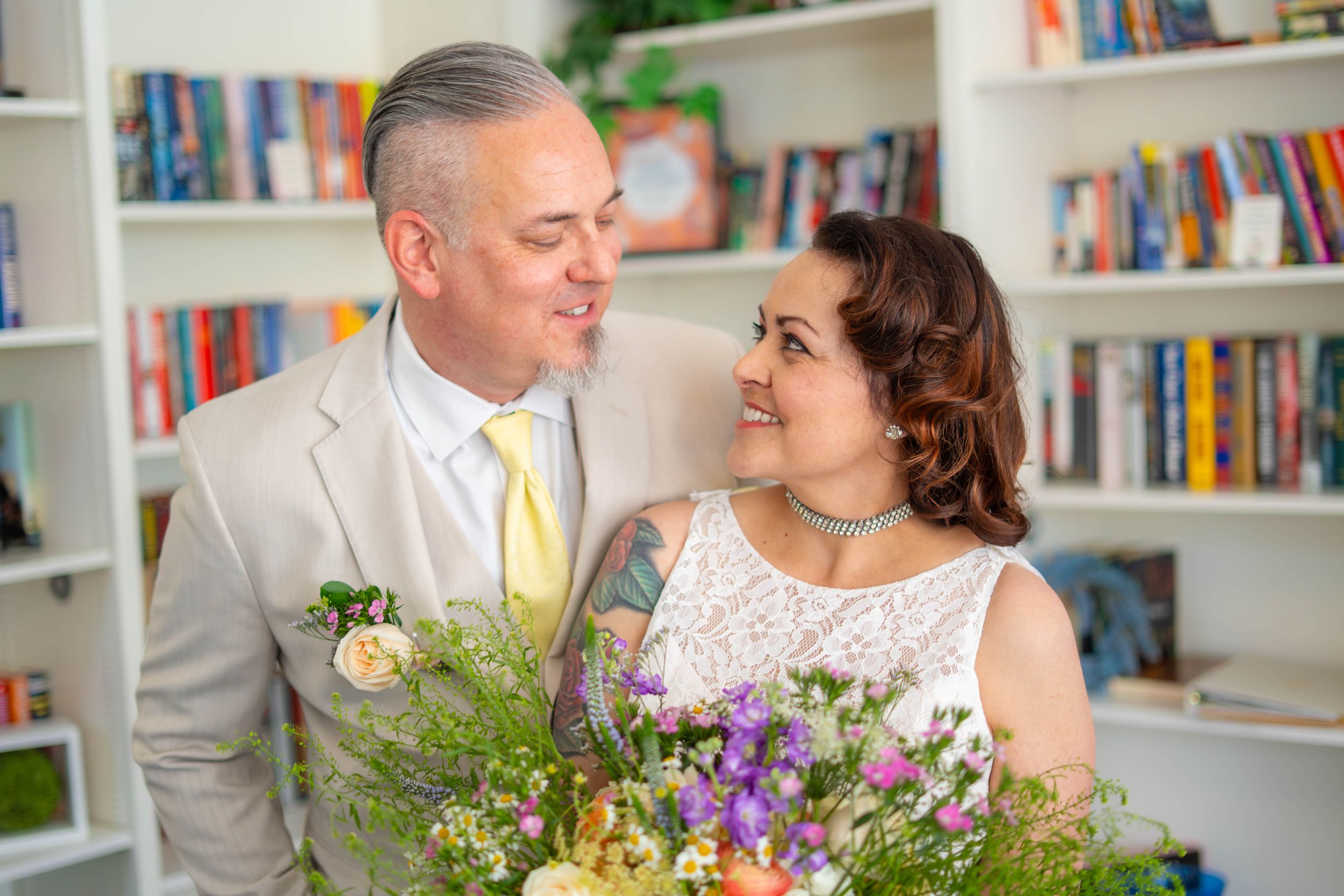 Couple in wedding attire smiling at each other holding colorful bouquets in front of bookshelves.  Quad Cities Wedding Photographer