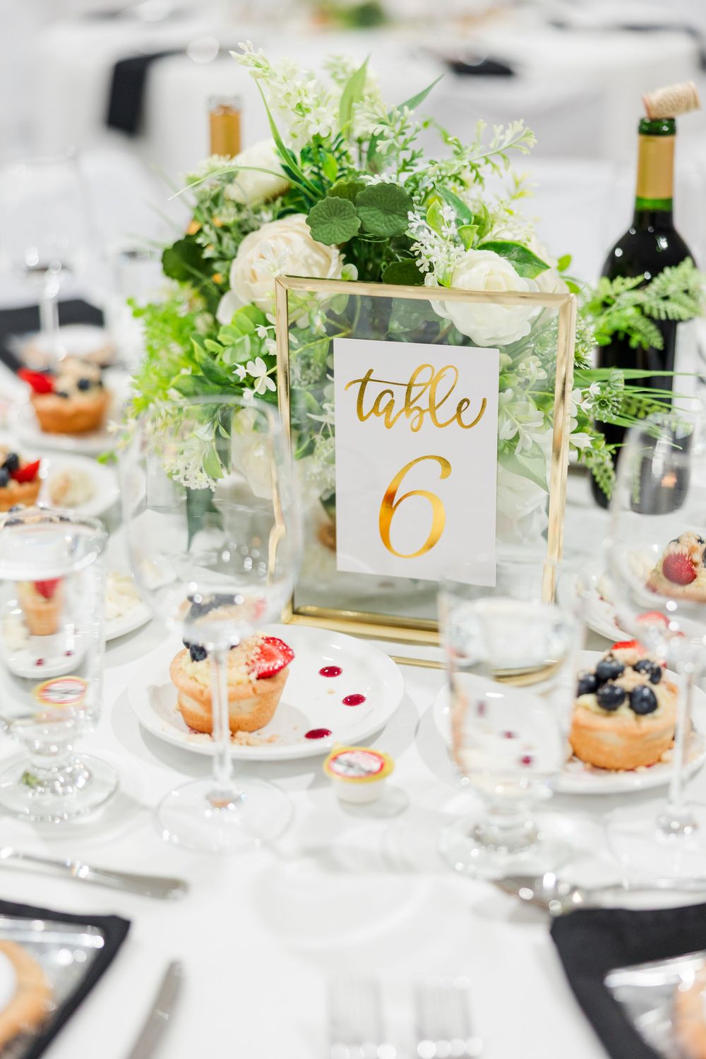 Elegant wedding reception table 6 with gold frame number sign, white floral centerpiece, and desserts on white linen.