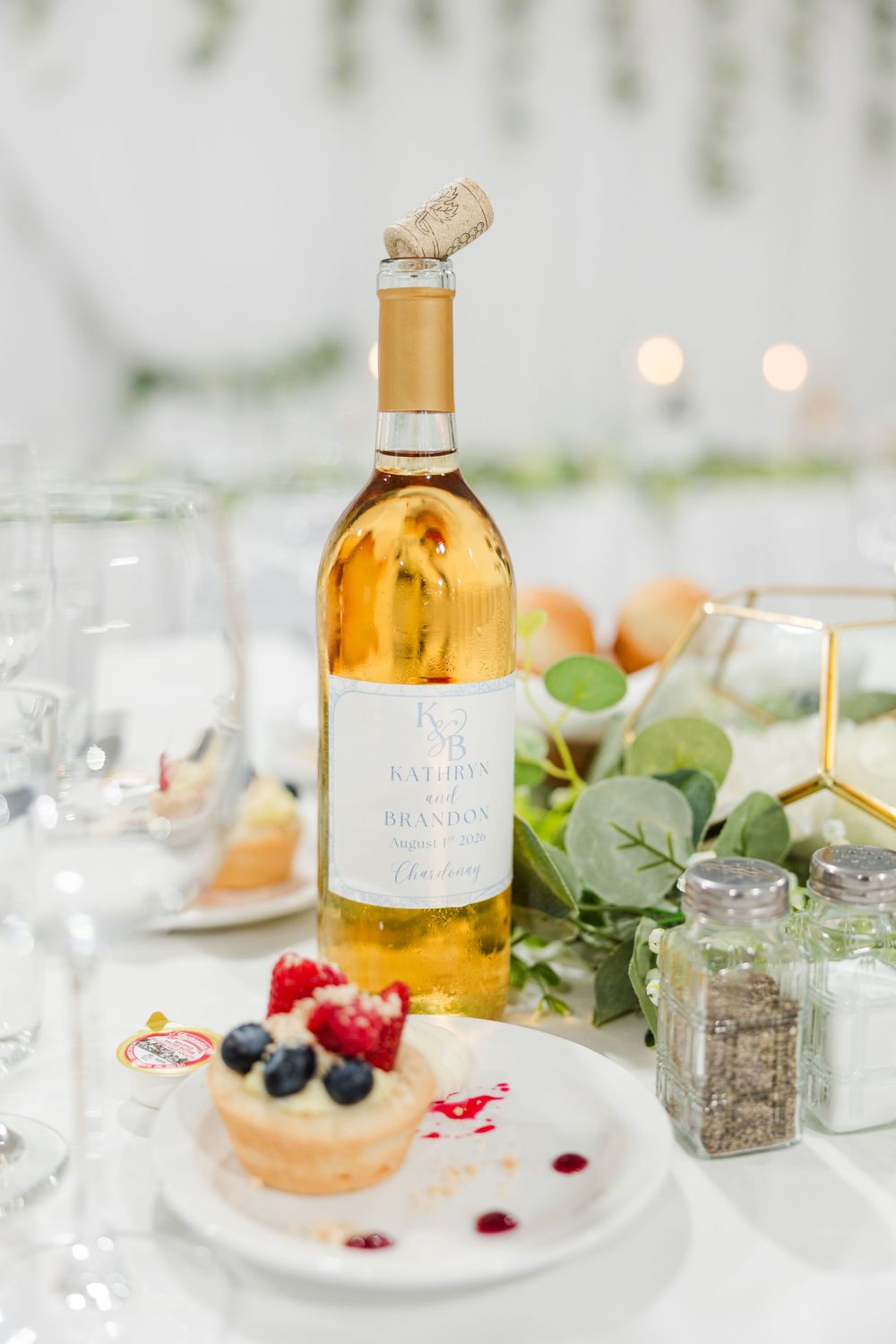 Elegant wedding table with personalized wine bottle, berry tart dessert, greenery, and glassware at a reception.