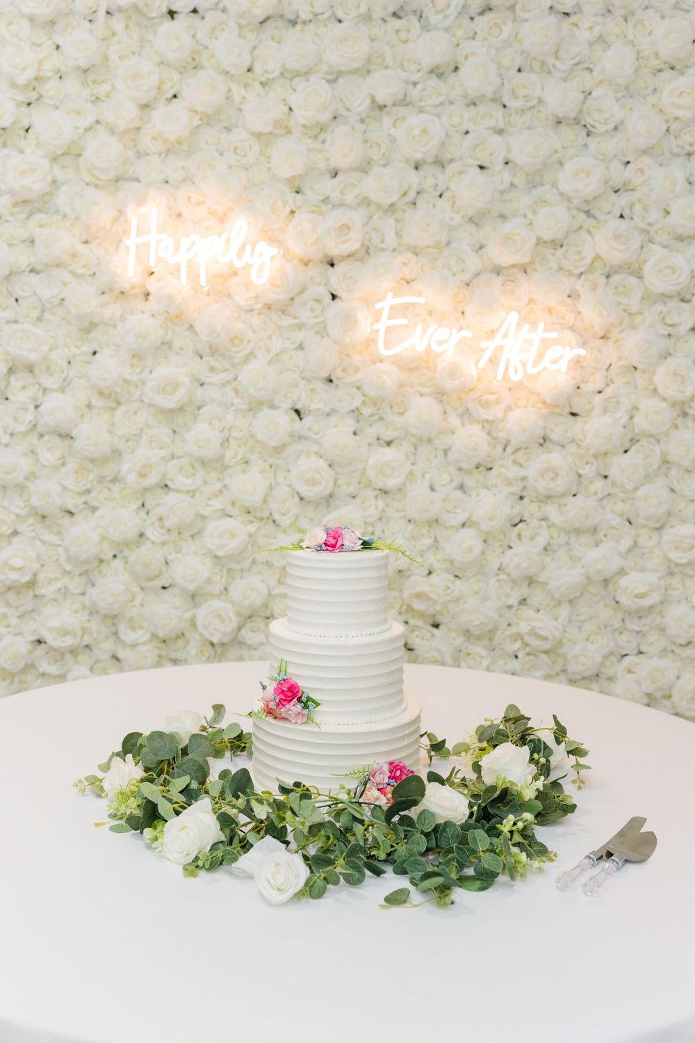 Three-tier white wedding cake on floral table with white rose wall backdrop and neon Happily Ever After sign.