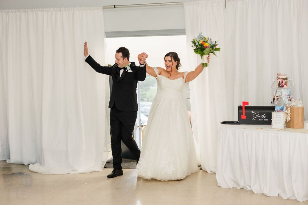 Bride and groom celebrate their grand entrance at wedding reception, arms raised in joy, white drapes backdrop.