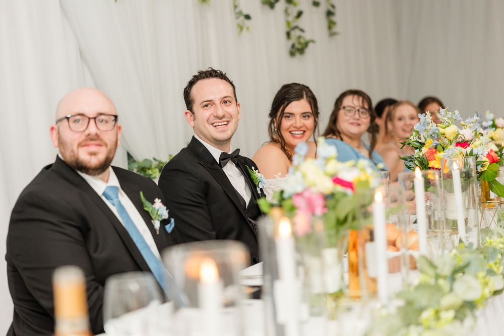 Wedding party smiles at head table during reception ceremony indoors.