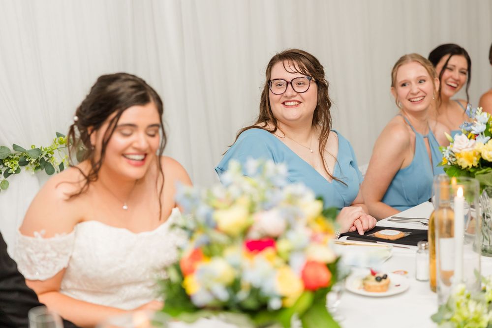 Bride smiling at wedding reception table with bridesmaids in blue dresses and colorful floral centerpieces.