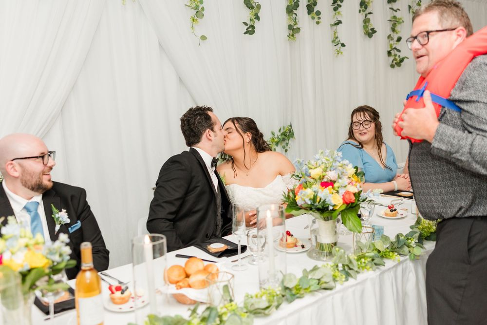 Bride and groom share a kiss at wedding reception head table while guests smile and laugh nearby.
