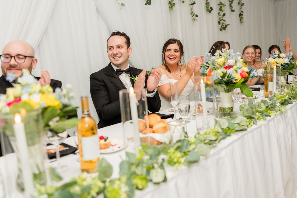 Bride and groom laughing and clapping at wedding reception head table decorated with greenery and colorful flowers.