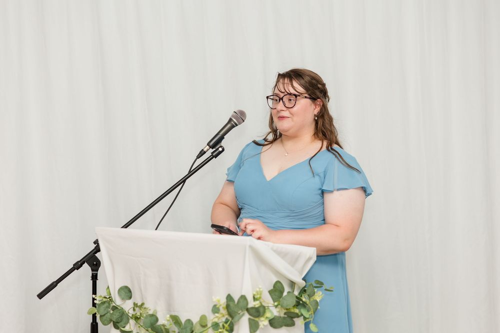 Woman in blue dress speaking at a podium with microphone at a formal event with white drape backdrop.