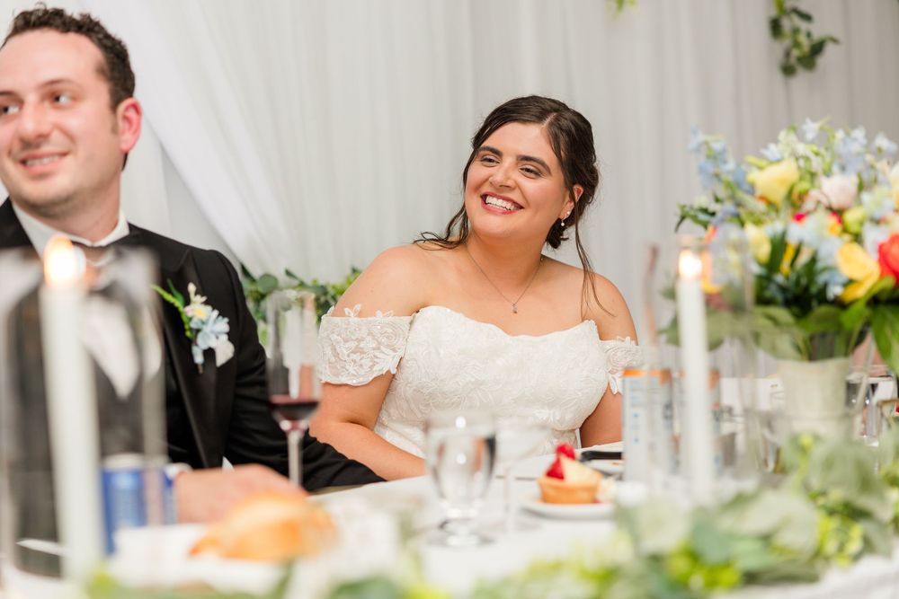 Smiling bride in white off-shoulder gown seated at wedding reception table with groom in black tuxedo.