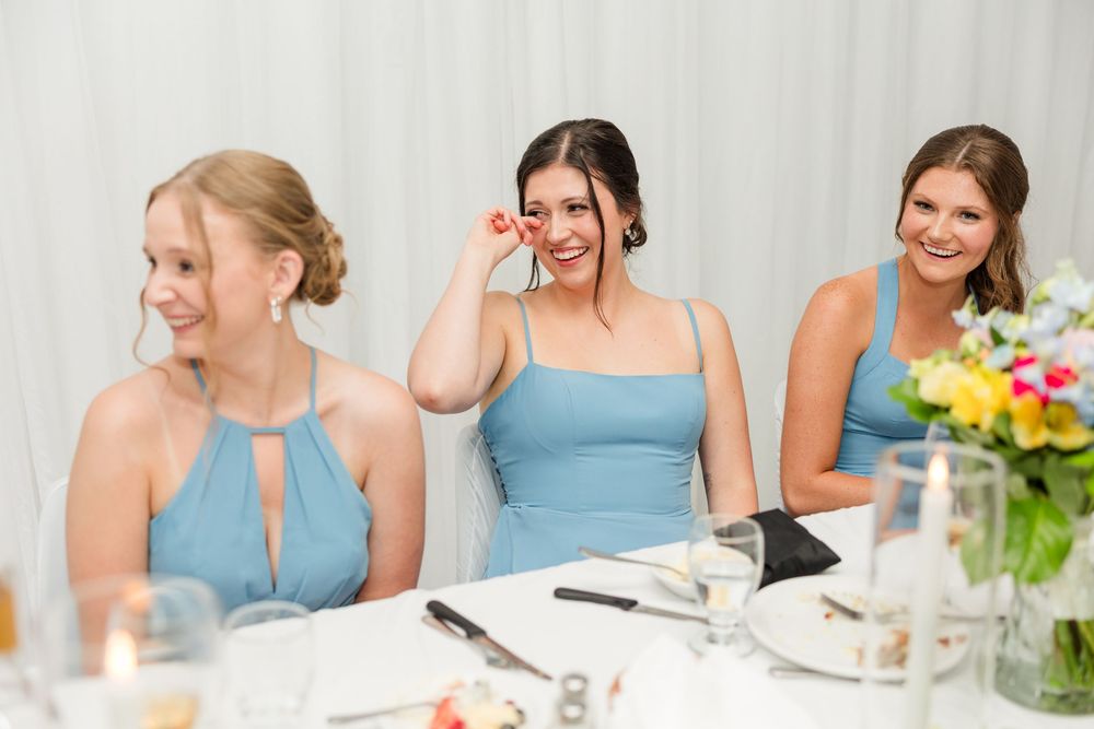 Three bridesmaids in blue dresses laughing and smiling at a wedding reception table with flowers.