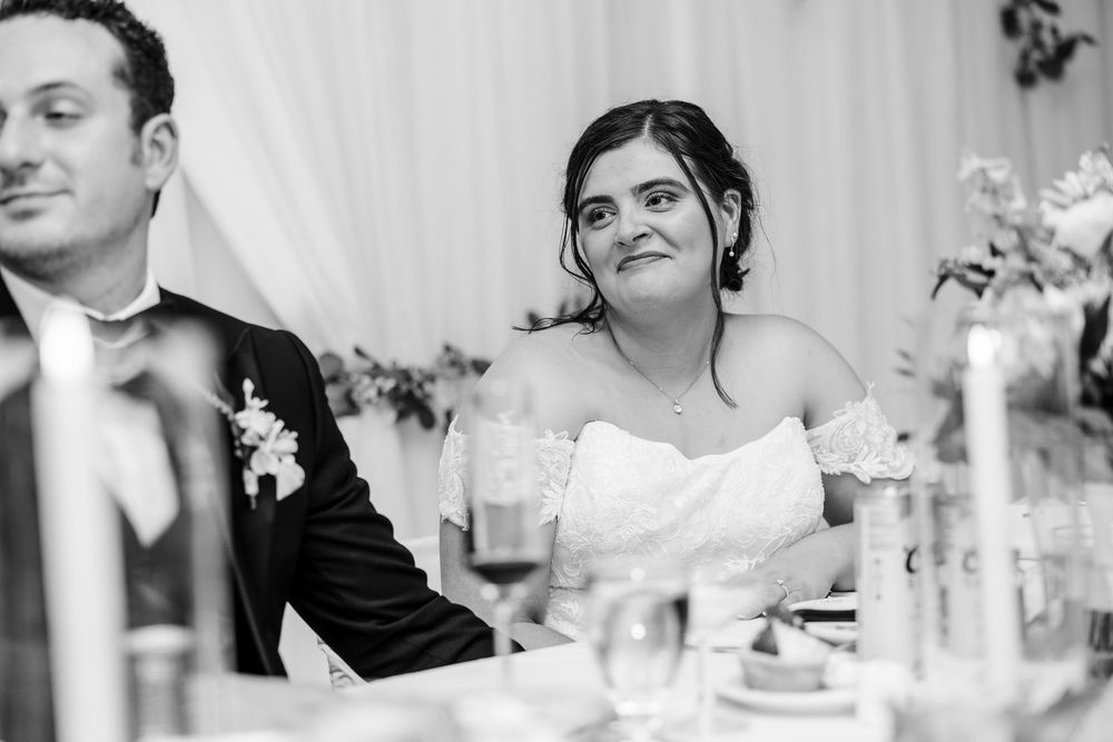 Bride in off-shoulder gown smiling during wedding reception speech, black and white photo.