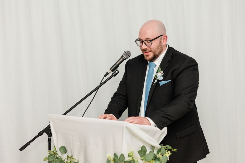 Bald man in black suit speaking at podium with microphone during wedding reception.