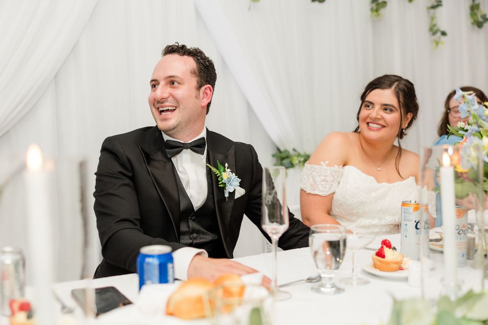 Smiling bride and groom in formal attire laughing together at wedding reception head table.