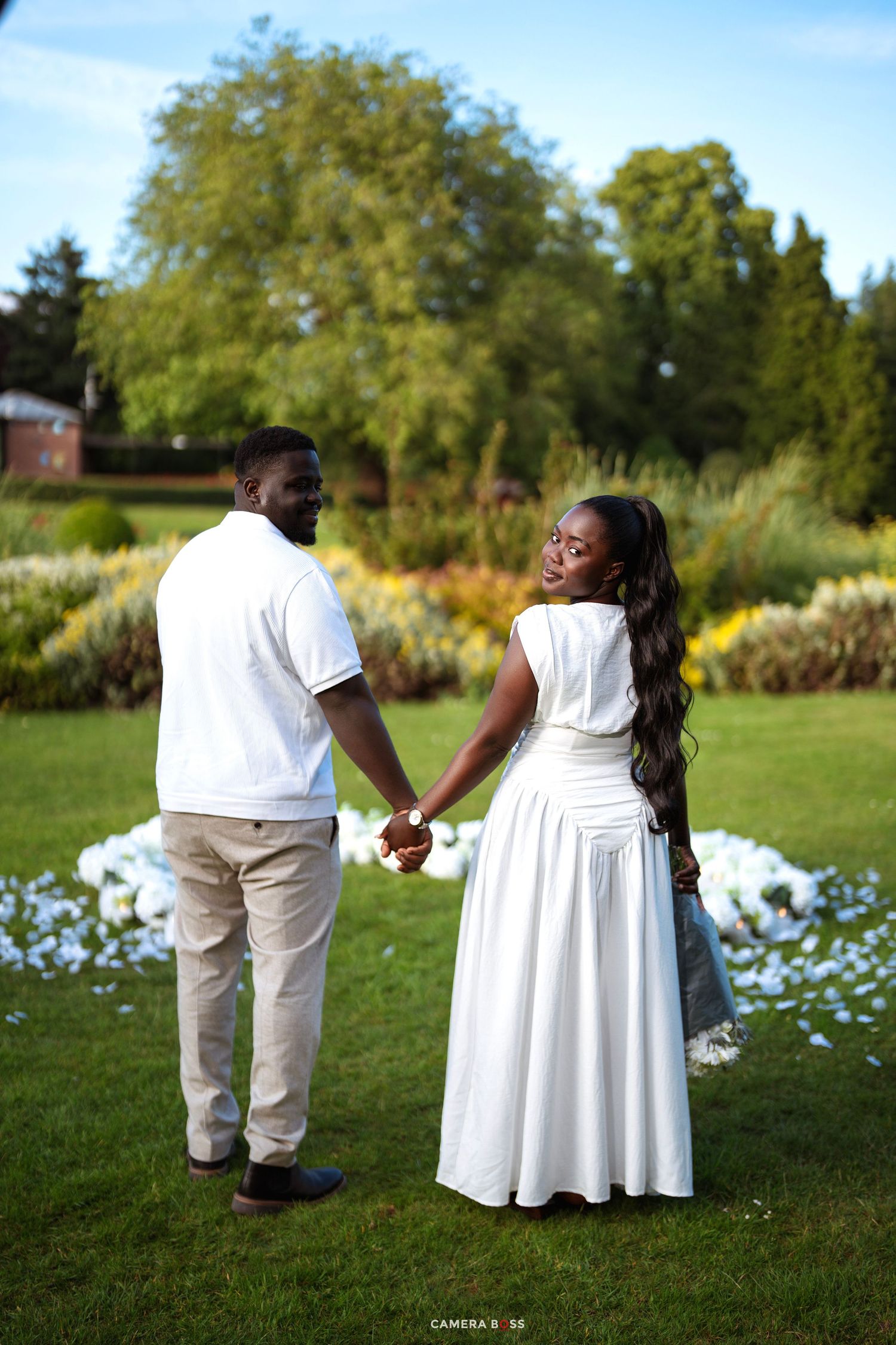Couple in white outfits holding hands in a lush green garden, woman looking back and smiling.