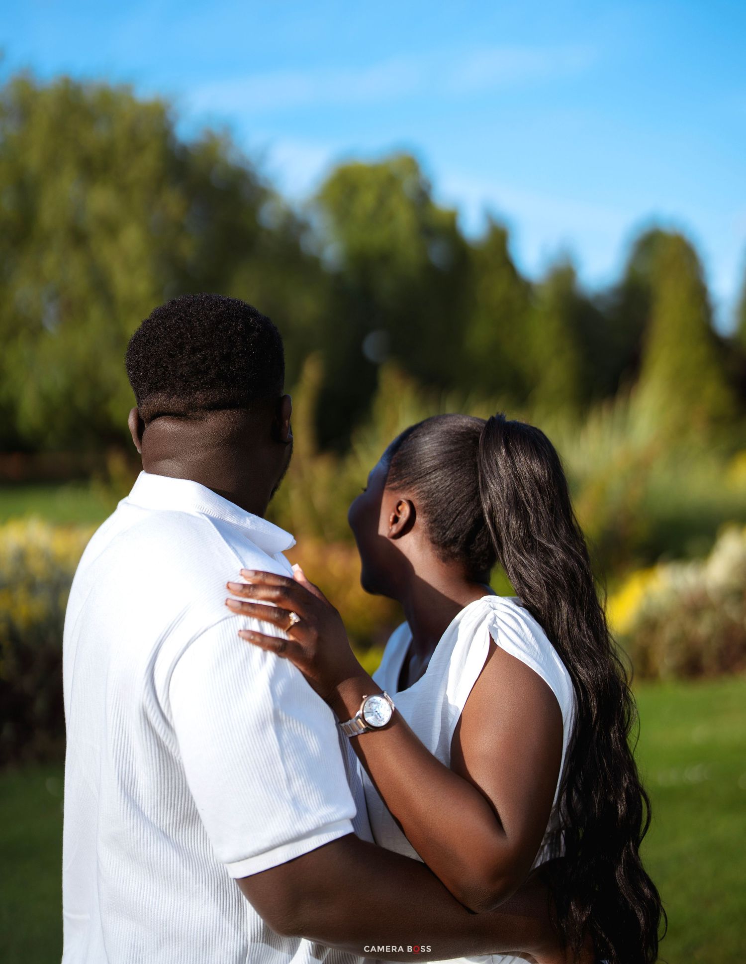 A couple in white outfits embraces outdoors in a lush green garden on a sunny day.