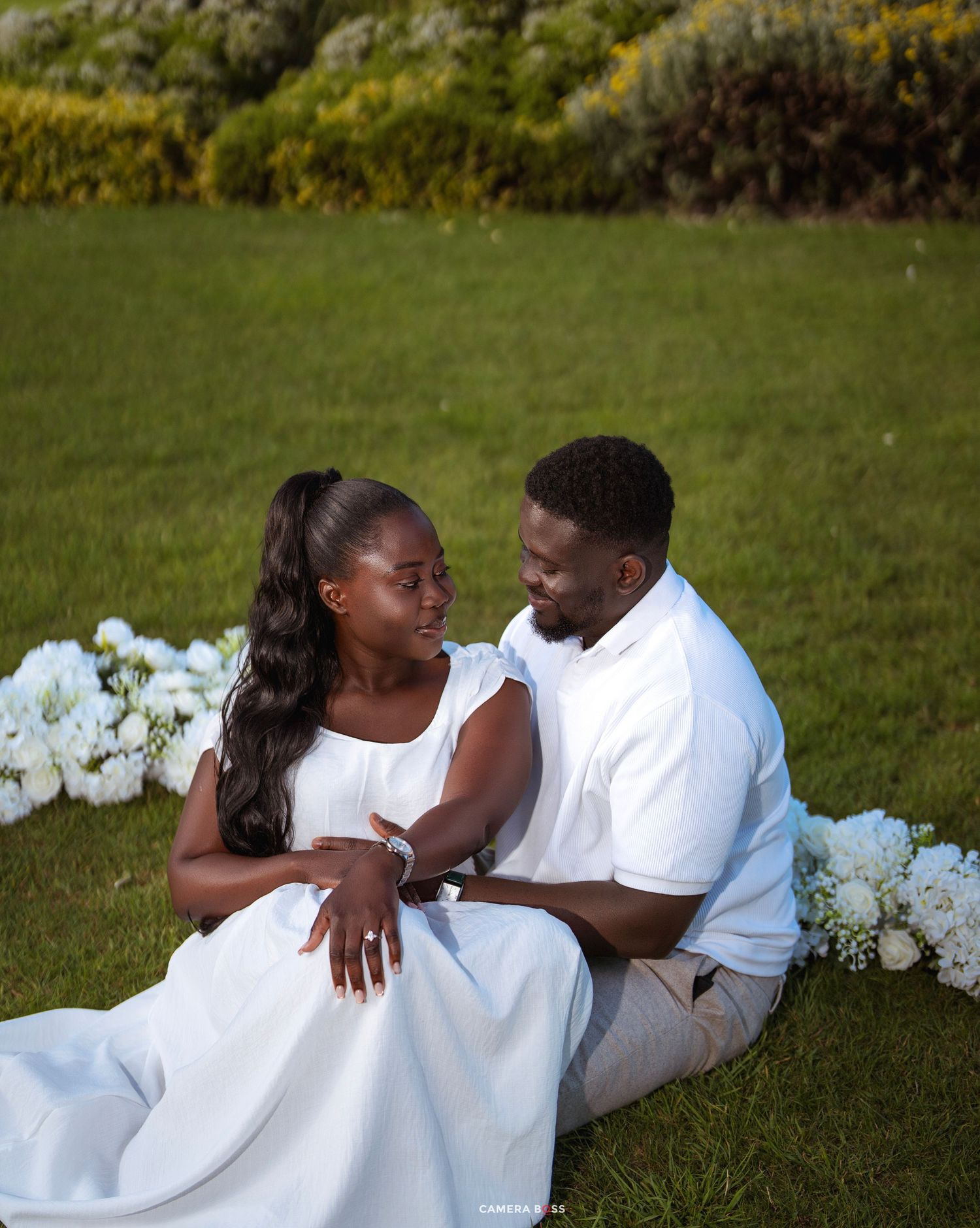 Couple in white outfits sitting on green grass surrounded by white hydrangeas during an outdoor engagement photo session.