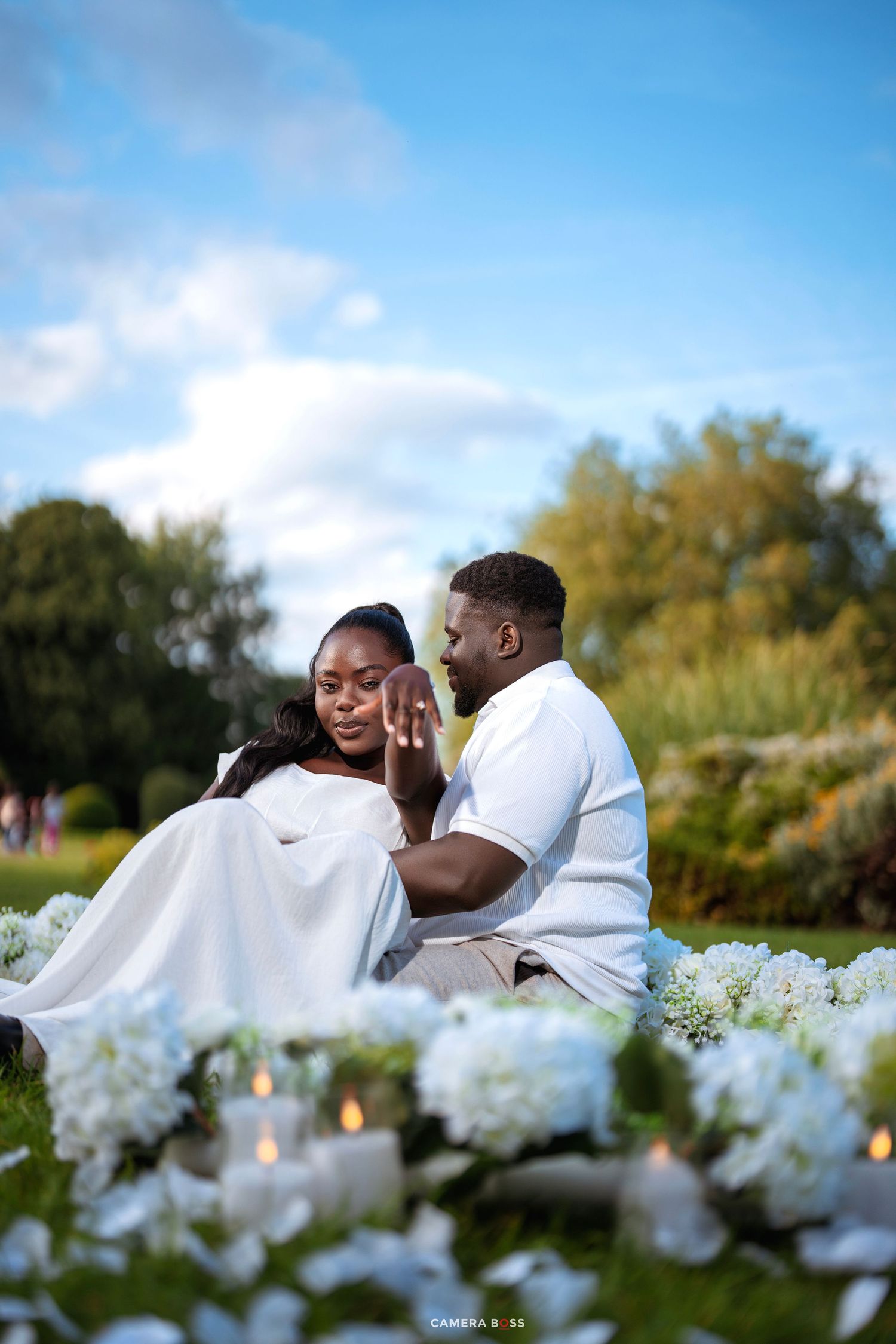 Couple in white outfits sharing a romantic outdoor proposal moment surrounded by white flowers and candles on green grass.