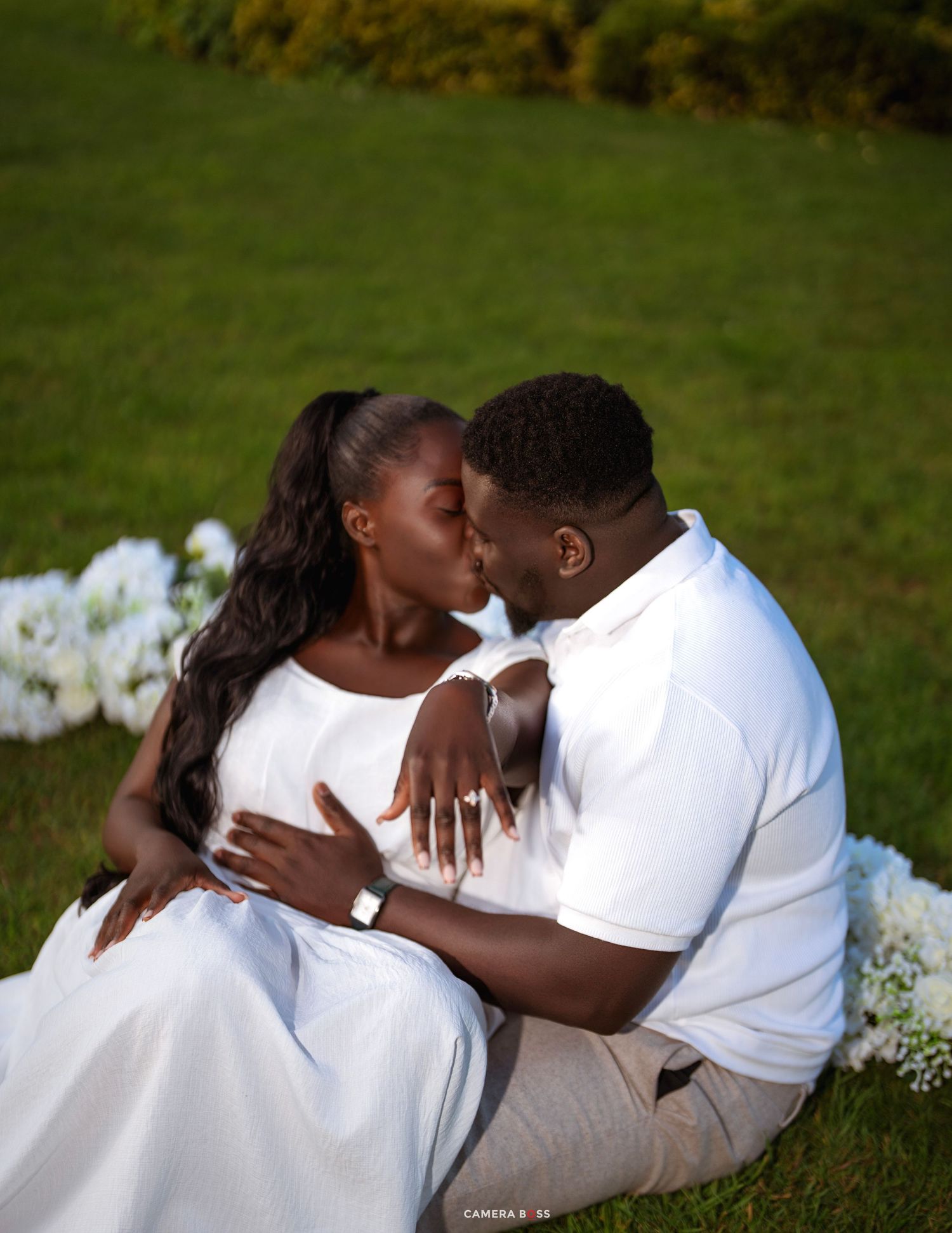 A couple sharing a romantic kiss while sitting on green grass, dressed in white outfits, with white flowers nearby.