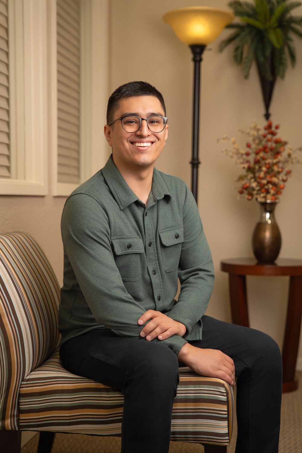 Smiling young man in green shirt and glasses sitting relaxed in striped chair in warmly lit room.