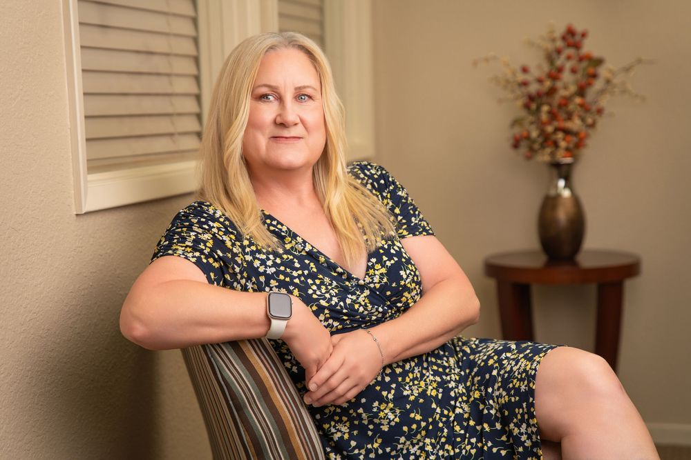 A blonde woman in a floral dress sits in a chair, wearing a smartwatch, posed in a warmly decorated room.