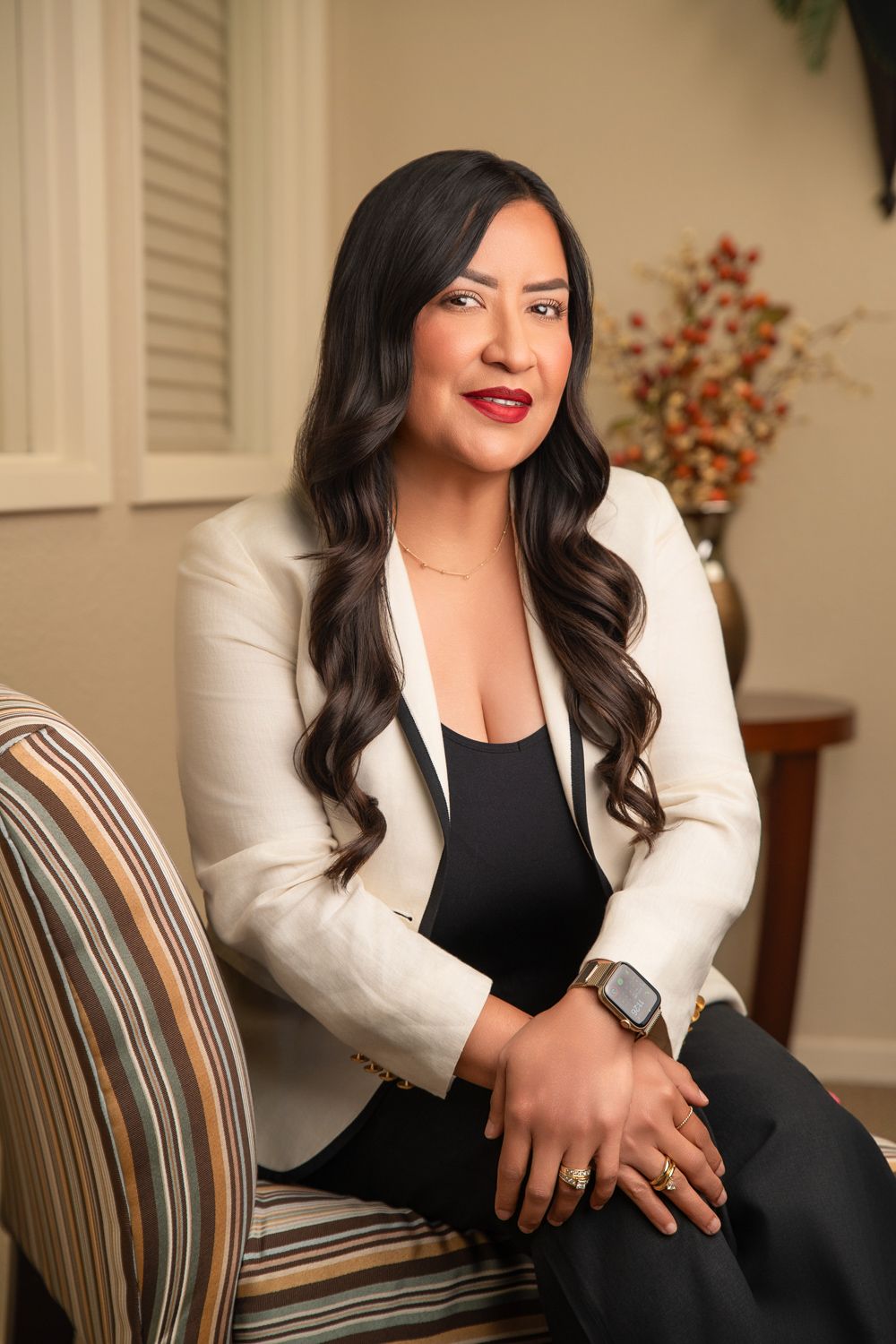 Professional woman in white blazer and black outfit seated elegantly in a striped chair in a warmly lit room.