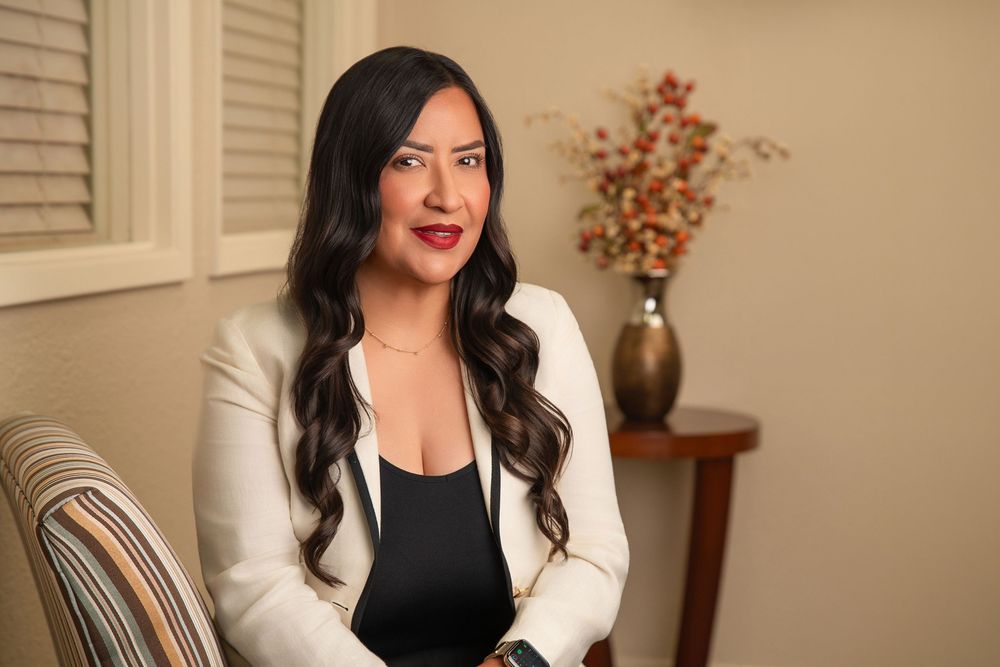 Professional woman with long dark hair wearing a white blazer and black top, seated in an elegant interior setting.