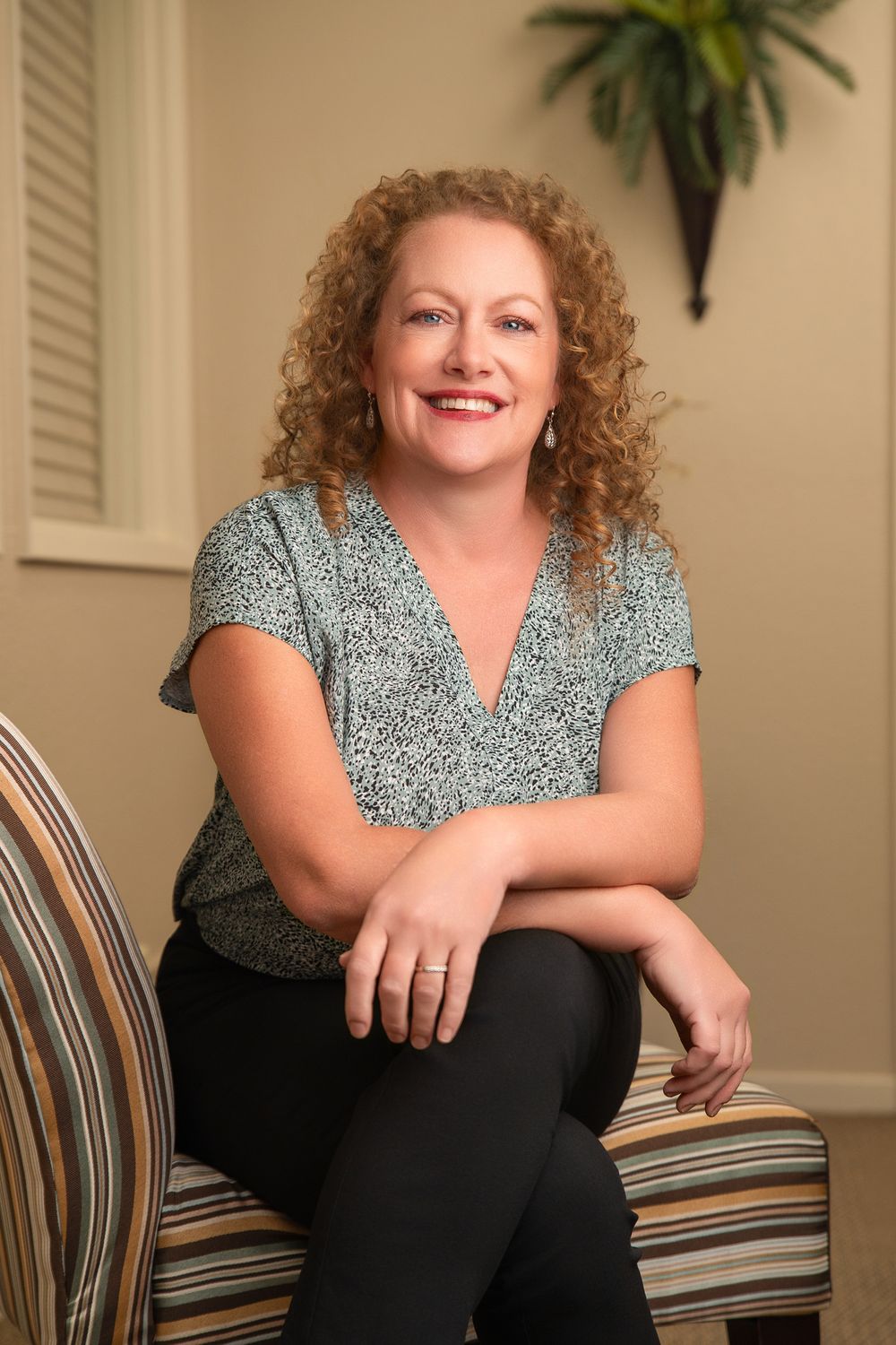 Smiling woman with curly red hair sits on striped chair wearing patterned top and black pants in warm interior setting.