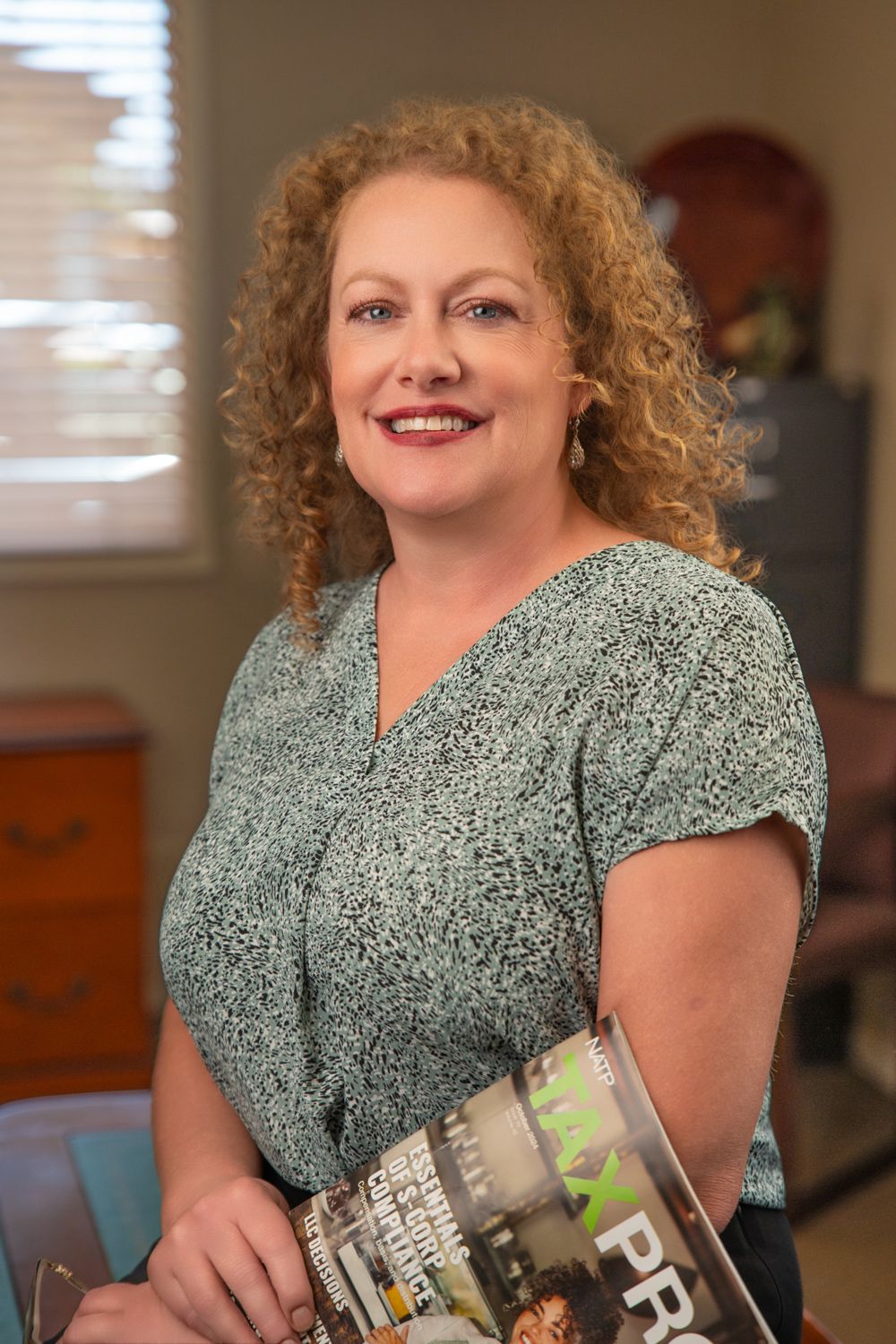 Smiling woman with curly red hair holding a magazine in a bright office, wearing a teal patterned blouse.