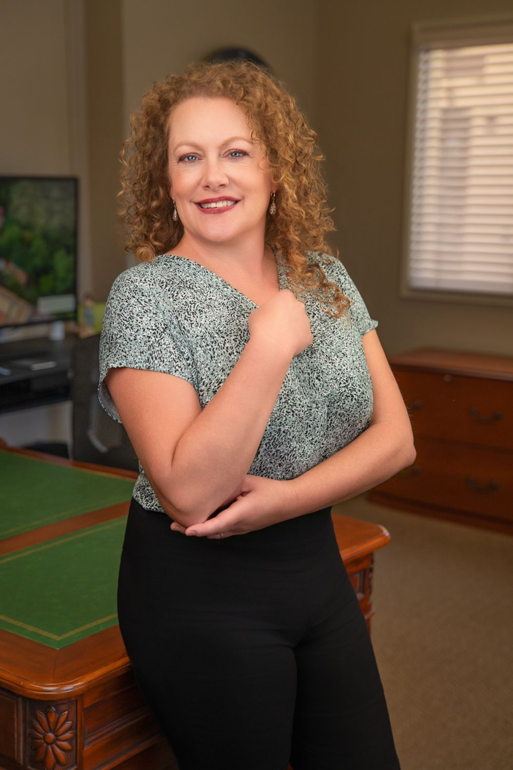Smiling woman with curly blonde hair wearing a green top and black pants, posing in a professional office setting.