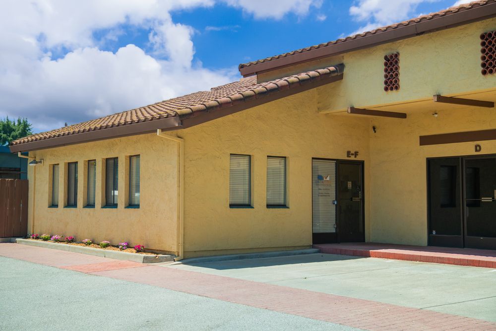 Yellow stucco school building with red tile roof, large windows, and covered entrance under partly cloudy blue sky.