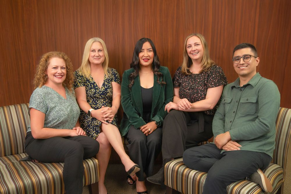 Five professional colleagues smiling and posing together on a couch in a modern office setting.