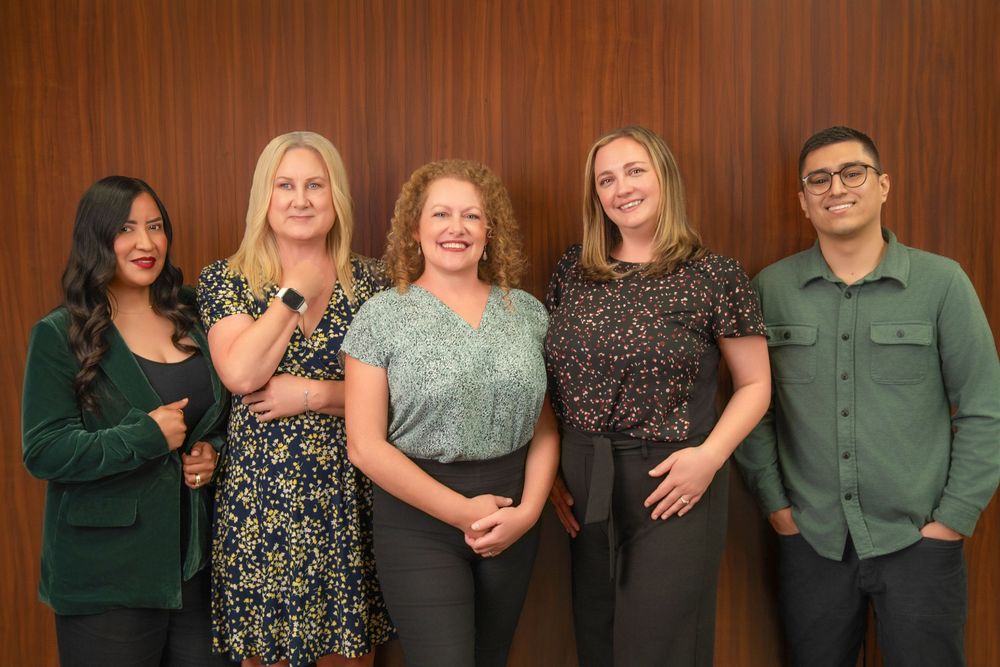Five professional colleagues smiling together in a corporate office setting with a wooden panel background.