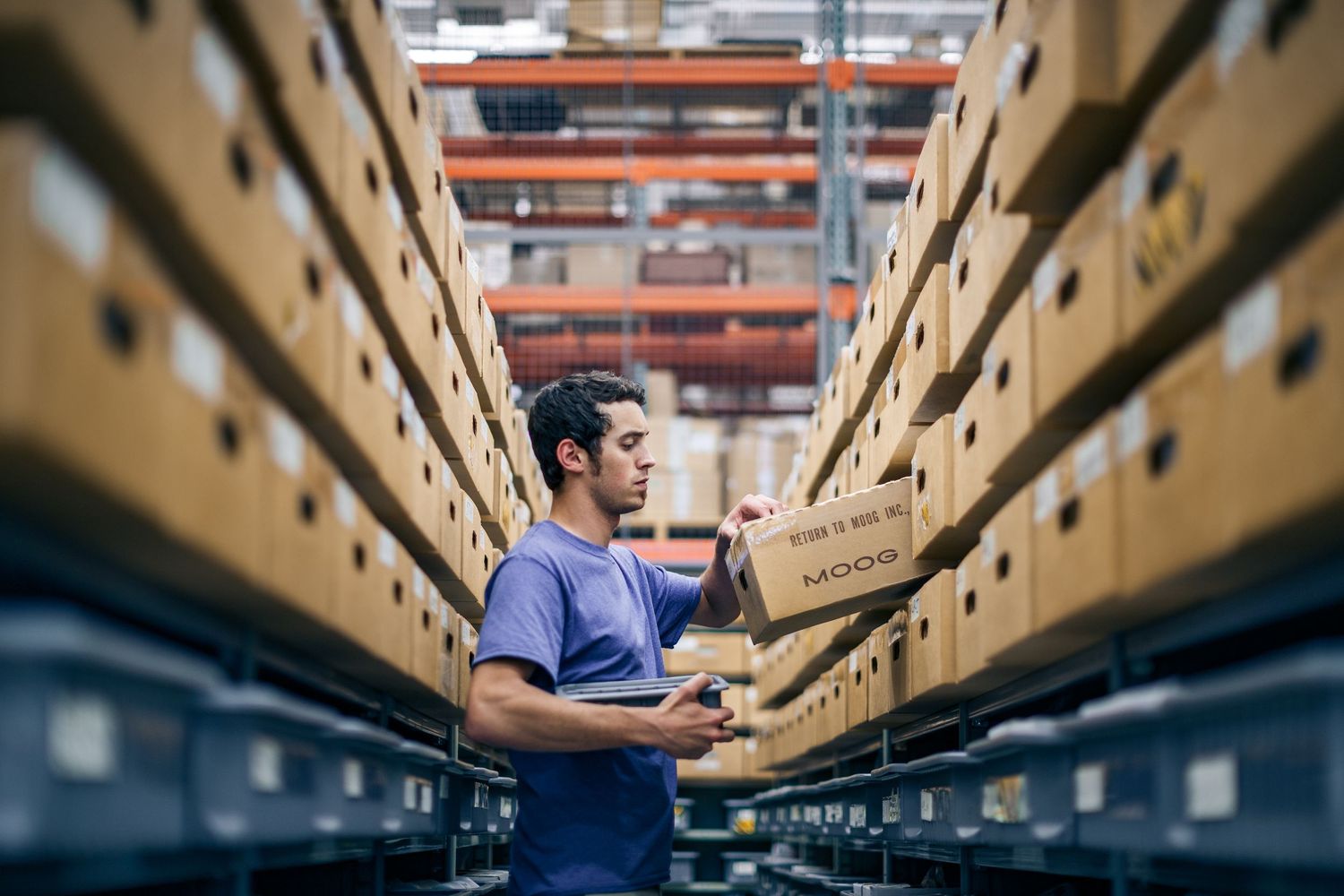 Warehouse worker in blue shirt scanning cardboard box on shelving rack in large distribution center.