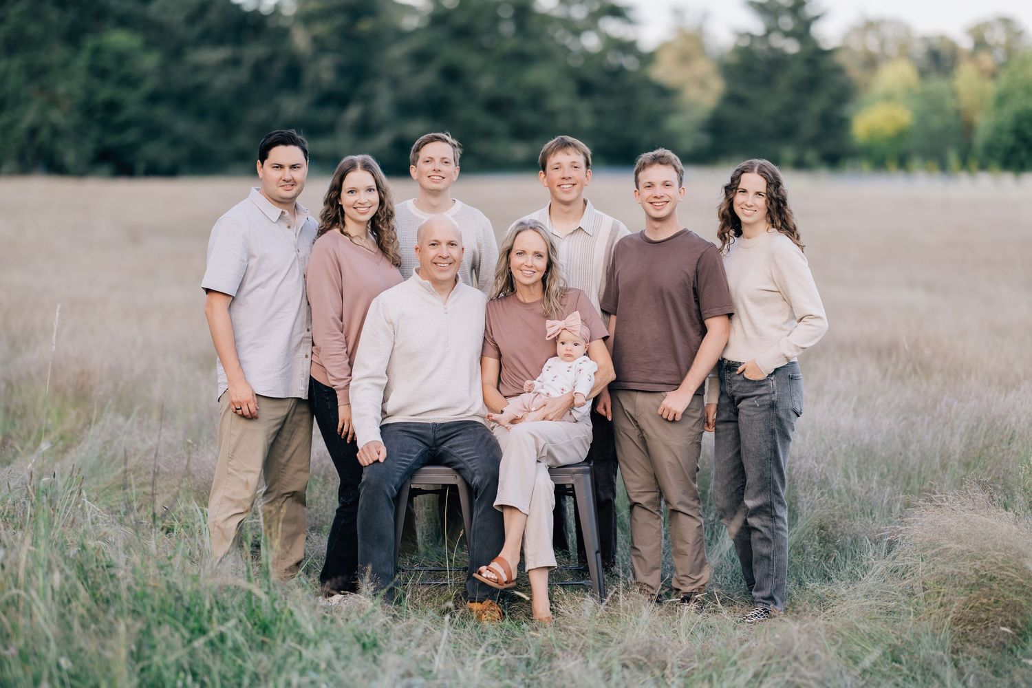 A large family of nine poses together in a golden field, wearing neutral tones for an outdoor portrait.
