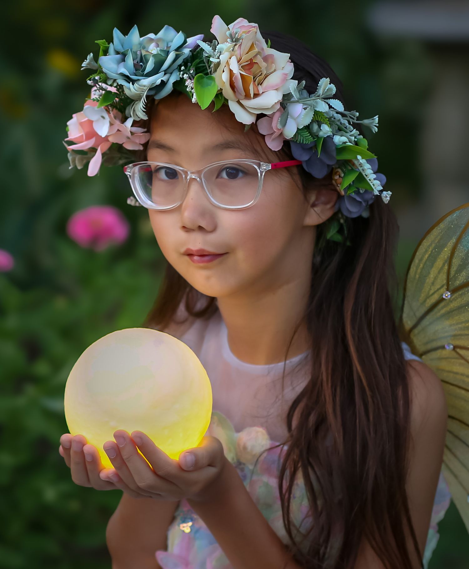 A young girl dressed as a fairy holds a glowing orb, wearing glasses and a floral crown in a garden setting.