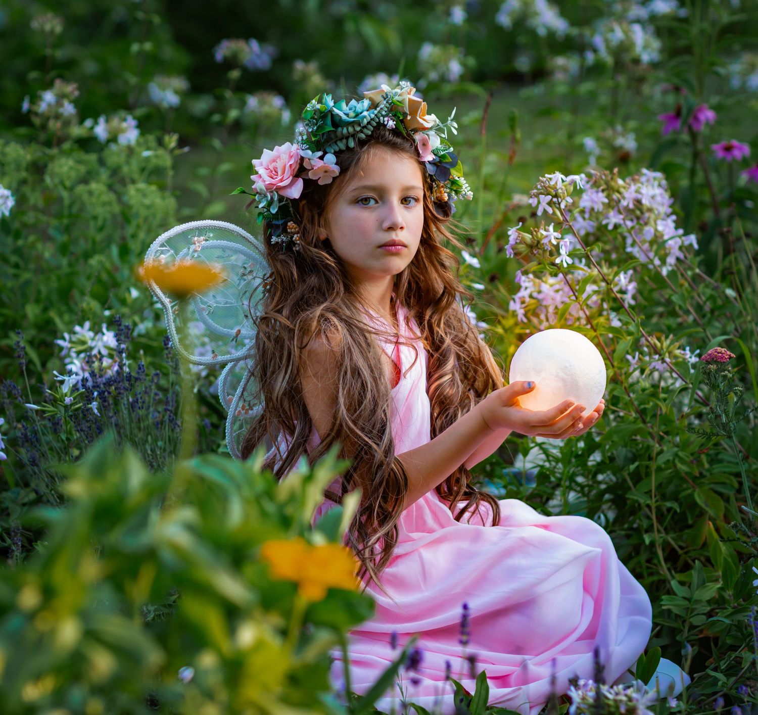 A young girl dressed as a fairy in a pink gown and floral crown holds a glowing orb among wildflowers.
