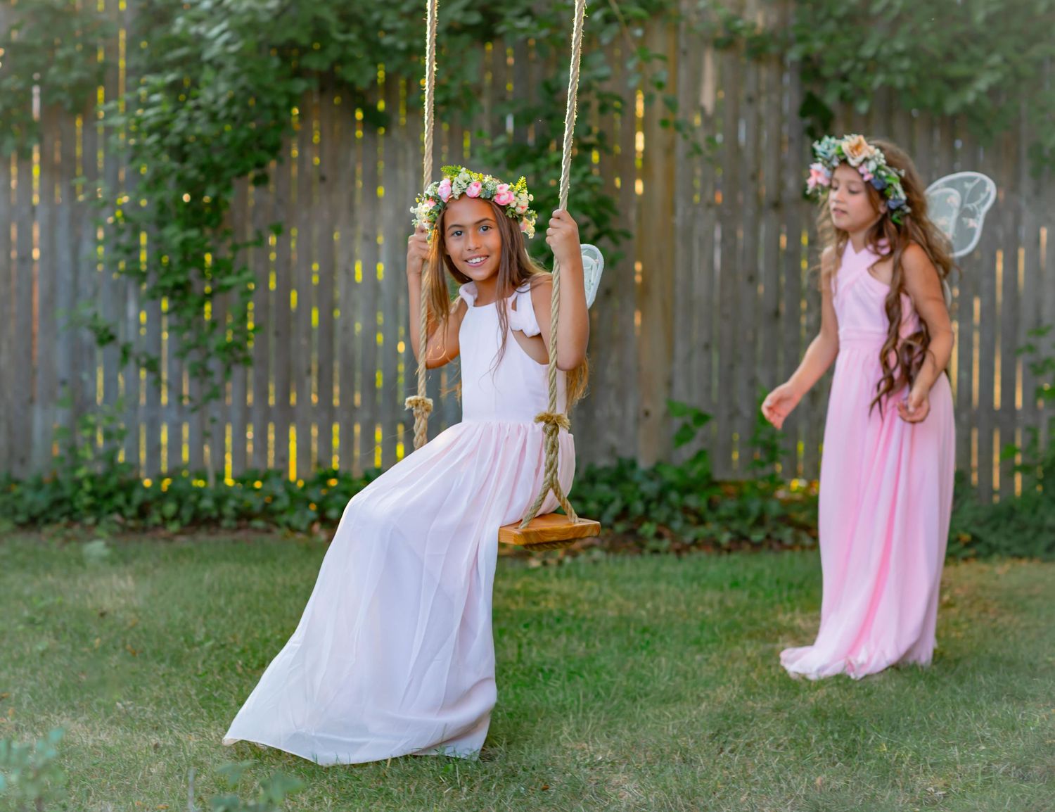 Two girls in white and pink dresses with flower crowns, one on a rope swing, in a lush backyard garden.